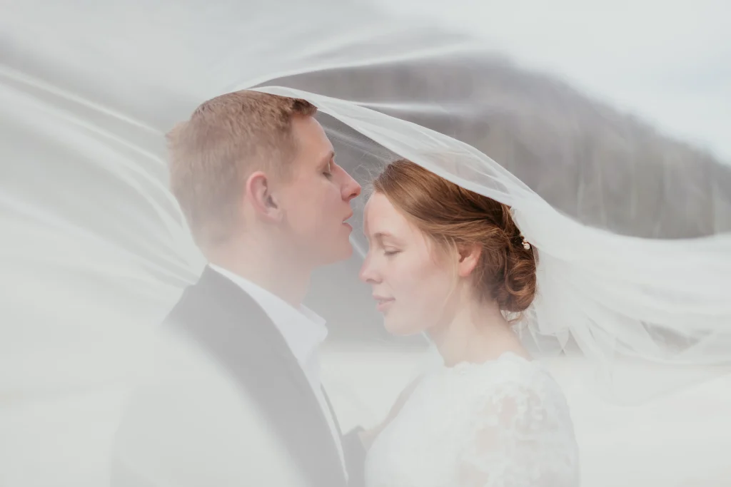"Bride and groom standing on the shore of Lake Bled, surrounded by the lake’s tranquil waters and the majestic Bled Castle in the distance."